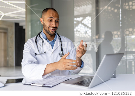 Smiling doctor with stethoscope uses laptop for virtual consultation in modern office. Engages in telemedicine call, representing efficient healthcare technology and professional communication. 121085132