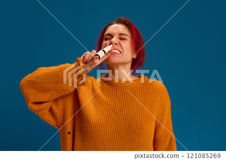 Portrait of young woman feels sick because of sneezing and spraying her nose to remove symptoms against dark blue background. 121085269