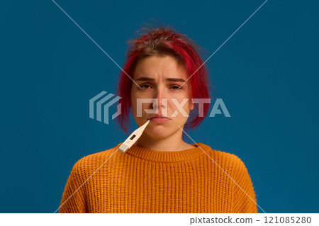 Portrait of sad, depressed young woman with thermometer in mouth measuring temperature of body against dark blue background. Portrait of sad, depressed young woman with thermometer in mouth measuring temperature of body against dark blue background. 121085280