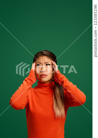 Young Asian woman holding hands on head as she feels pain dressed against green studio background. Woman feels badly. 121085296