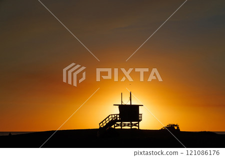 Los Angeles beach at sunset, featuring a lifeguard tower silhouette against a glowing orange and purple horizon 121086176