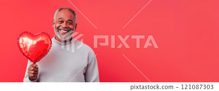 Cheerful elderly African American man in light sweater holding heart-shaped balloon on red background. Valentine's Day banner 121087303