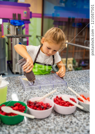 Little girl learning how to cook in a cooking class. Handmade dessert. Making chocolate candy, Child filling heart shaped mold with chocolate Little girl learning how to cook in a cooking class. Handmade dessert. Making chocolate candy, Child filling heart shaped mold with chocolate 121087501