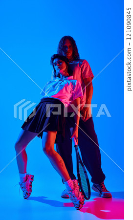 Portrait of stylish young man and woman wearing trendy clothes and posing with tennis racket against blue studio background in neon light 121090845