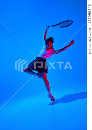 Dynamic image of young sportive woman in shirt and skirt, posing in motion with tennis racket against blue studio background in neon light 121090846