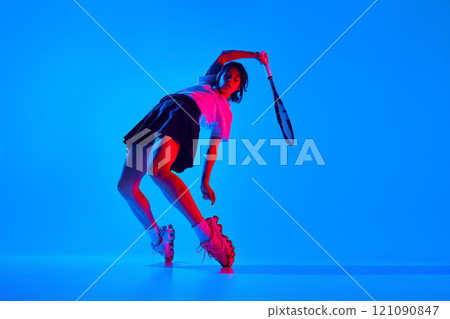 Dynamic image of young sportive woman in shirt and skirt, posing in motion with tennis racket against blue studio background in neon light 121090847