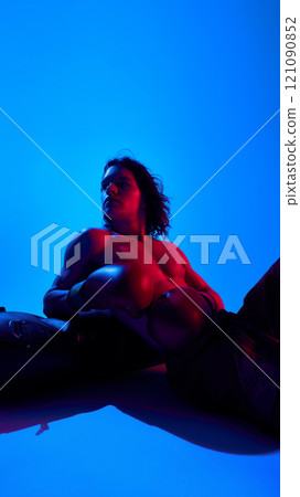 Handsome young shirtless man with muscular body. in boxing gloves resting punching bag against blue studio background in neon light 121090852