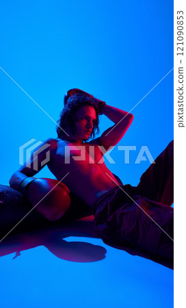 Handsome young shirtless man with muscular body. in boxing gloves resting punching bag against blue studio background in neon light 121090853