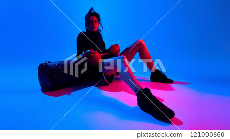 Stylish and confident young woman in boxing gloves sitting on punching bag after intense training against blue studio background in neon light 121090860