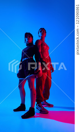 Low angle portrait of shirtless man and woman in sportswear, wearing boxing gloves, posing against blue studio background in neon light. Full-length 121090861