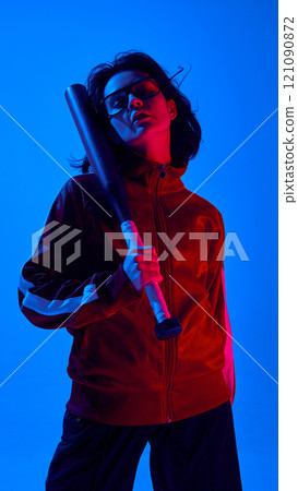 Portrait of confident young woman in casual black glasses, red sportive jacket, stranding with baseball bat against blue studio background in neon light 121090872