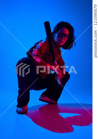Stylish confident young woman in glasses and sportive attire, sitting and posing with baseball bat against blue studio background in neon light Stylish confident young woman in glasses and sportive attire, sitting and posing with baseball bat against blue studio background in neon light 121090879