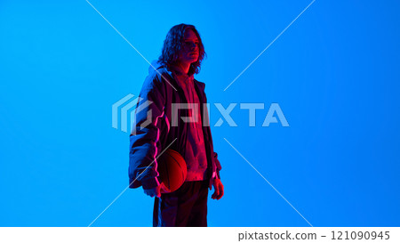 Side view portrait of handsome young man with long curly hair, wearing oversized sportive clothes standing with basketball against blue studio background in neon light 121090945