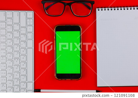 A phone with a green chroma key screen is lying on a red background, to the right of it is a white notebook with black springs on top, black glasses with transparent glasses are on top, a white A phone with a green chroma key screen is lying on a red background, to the right of it is a white notebook with black springs on top, black glasses with transparent glasses are on top, a white 121091108