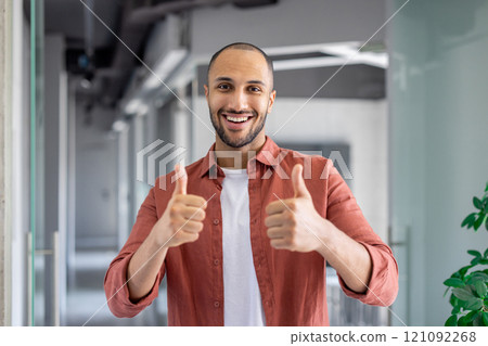 Smiling young man in a casual shirt giving two thumbs up indoors, symbolizing positivity and encouragement. His cheerful expression conveys optimism, enthusiasm, and confidence in workspace Smiling young man in a casual shirt giving two thumbs up indoors, symbolizing positivity and encouragement. His cheerful expression conveys optimism, enthusiasm, and confidence in workspace 121092268