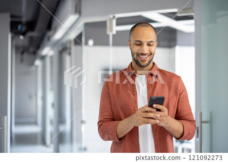 Confident young professional enjoying moment while checking smartphone in modern office hallway. Casual yet focused, he engages with tasks using phone, embodying work-life balance. 121092273