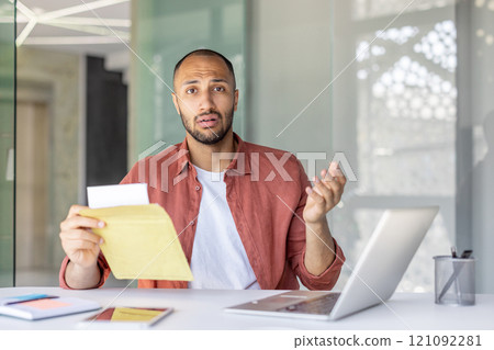 A confused office worker holds an envelope while sitting at a desk with a laptop. His expression suggests surprise or misunderstanding. Various devices and papers are on the desk. A confused office worker holds an envelope while sitting at a desk with a laptop. His expression suggests surprise or misunderstanding. Various devices and papers are on the desk. 121092281