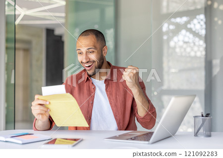 A joyful man celebrates good news at his office desk, displaying excitement and success. He holds an envelope and works on his laptop, evoking feelings of achievement. A joyful man celebrates good news at his office desk, displaying excitement and success. He holds an envelope and works on his laptop, evoking feelings of achievement. 121092283