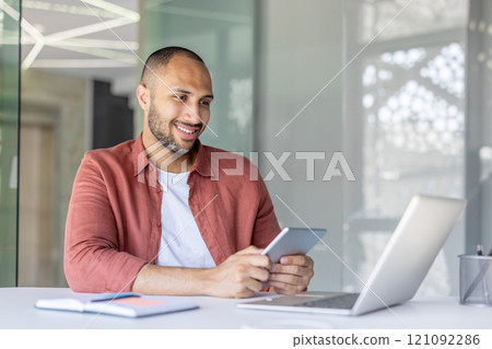 A smiling office worker interacts with a tablet and laptop in a contemporary workspace. The atmosphere is professional and innovative, highlighting the use of technology in a modern office setting. A smiling office worker interacts with a tablet and laptop in a contemporary workspace. The atmosphere is professional and innovative, highlighting the use of technology in a modern office setting. 121092286