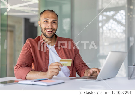 Smiling man using a laptop and holding a credit card, suggesting online shopping. The setup indicates a modern office environment, promoting digital technology and financial transactions Smiling man using a laptop and holding a credit card, suggesting online shopping. The setup indicates a modern office environment, promoting digital technology and financial transactions 121092291