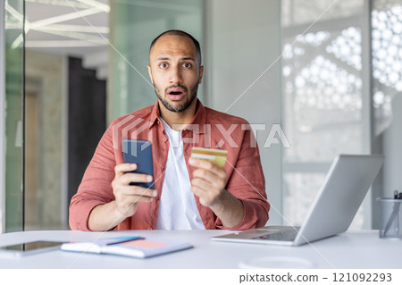 A surprised man is holding a smartphone and a credit card while sitting at a desk. He appears shocked, possibly due to unexpected news or online transaction issues. 121092293