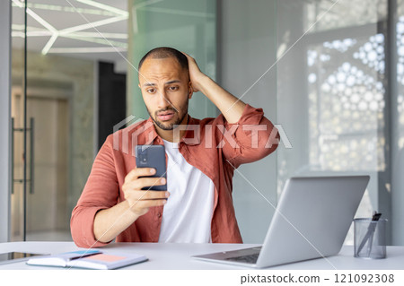 Concerned man holds smartphone and looks worried while working in office. Laptop and notebook on desk suggest business or remote work setting. Casual clothing adds relaxed vibe. 121092308