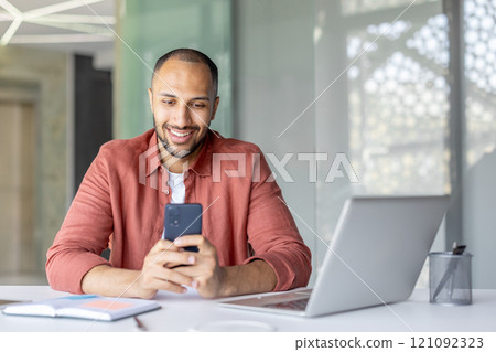 A cheerful male professional interacts with his smartphone while multitasking, working at his laptop. His positive demeanor embodies efficiency, communication, and productivity in work environment. A cheerful male professional interacts with his smartphone while multitasking, working at his laptop. His positive demeanor embodies efficiency, communication, and productivity in work environment. 121092323