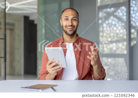 A cheerful man is using a tablet while engaging in a lively discussion at the office. His confident expression and professional attire suggest a productive work conversation. A cheerful man is using a tablet while engaging in a lively discussion at the office. His confident expression and professional attire suggest a productive work conversation. 121092346