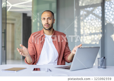 Portrait of upset and disappointed businessman, man looking displeasedly at camera while sitting inside office with laptop at workplace. Portrait of upset and disappointed businessman, man looking displeasedly at camera while sitting inside office with laptop at workplace. 121092359