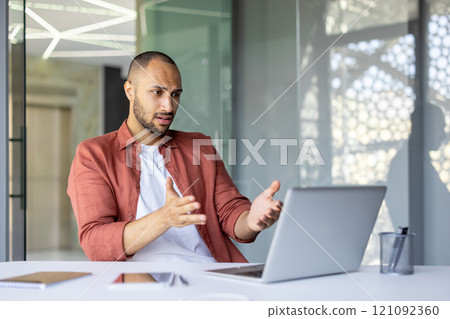 Young professional using laptop gesturing expressively during online meeting. Confused and frustrated looks, showing concern. Modern office setting indicates technology and communication challenges. 121092360