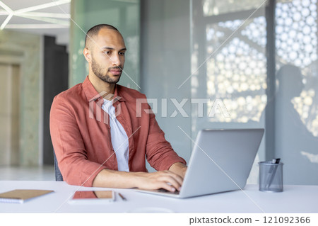 Confident businessman working diligently on a laptop in a bright, modern office. The professional setting includes essentials like a notebook, pen, and tablet, emphasizing focus and productivity. Confident businessman working diligently on a laptop in a bright, modern office. The professional setting includes essentials like a notebook, pen, and tablet, emphasizing focus and productivity. 121092366