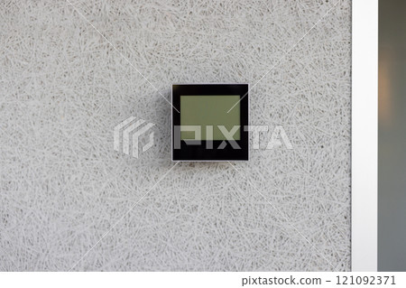 Electronic thermostat mounted on white wall displaying current indoor temperature. Device is sleek, modern, and essential for climate control in a minimalist environment. Electronic thermostat mounted on white wall displaying current indoor temperature. Device is sleek, modern, and essential for climate control in a minimalist environment. 121092371