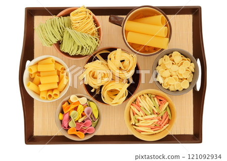 Varieties of multi-colored pasta. Types of uncooked pasta in bowls on a tray. Isolated on a white background. Storing different types of pasta at home. Top view. Varieties of multi-colored pasta. Types of uncooked pasta in bowls on a tray. Isolated on a white background. Storing different types of pasta at home. Top view. 121092934