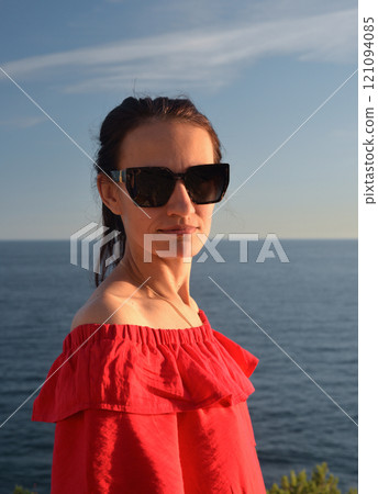 Portrait of a happy young woman in a red dress, against the background of the sea, looking at the camera with glasses. Portrait of a happy young woman in a red dress, against the background of the sea, looking at the camera with glasses. 121094085