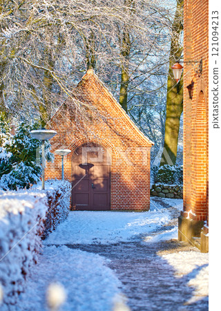 Exterior of an old church building with snow covered trees and pathway. Small chapel in snowy landscape with sun shining on brick wall. Countryside with European style architecture in winter scene 121094213