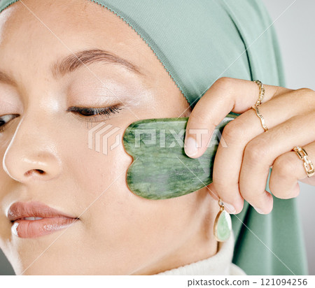 Muslim woman using a jade gua sha on face. Closeup headshot of an arab in a hijab sculpting face. Middle eastern woman using traditional chinese healing tool to aid lymph drainage, decrease puffiness Muslim woman using a jade gua sha on face. Closeup headshot of an arab in a hijab sculpting face. Middle eastern woman using traditional chinese healing tool to aid lymph drainage, decrease puffiness 121094256