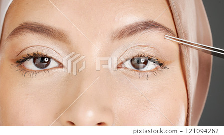 Closeup portrait of traditional muslim woman using tweezers to pluck her eyebrows. Half headshot of confident arab model wearing a hijab in a studio. Cultural middle eastern grooming her facial hair 121094262