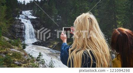 Woman, friends and camera with waterfall for sightseeing, picture or photography in nature. Female person, tourist or traveler with phone screen for memory, view or adventure by river in forest 121094298