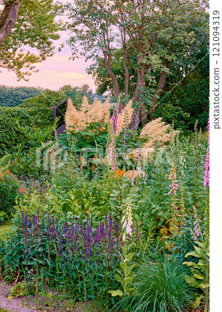 A landscape view of pink foxglove, yellow foxtail lily, orange tecoma, purple Delphinium, vines, and fern under a bright pink sky. A beautiful view of the forest with different tropical flora. 121094319