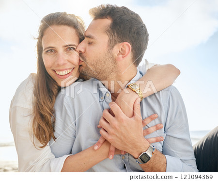 Beautiful middle aged woman being kissed on the cheek by her husband while sitting outdoors on the beach. Affectionate married couple hugging on a romantic date, showing love to his wife outside 121094369