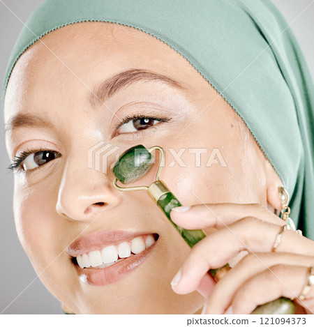 Muslim woman smiling and using a facial roller on her skin. Beauty model doing her morning skin care routine with a jade roller. Young female using a gua sha product for healthy smooth skin. Muslim woman smiling and using a facial roller on her skin. Beauty model doing her morning skin care routine with a jade roller. Young female using a gua sha product for healthy smooth skin. 121094373