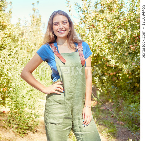 Portrait of a female farm worker standing on a fruit farm during harvest season. Young smiling farmer between fruit trees on a sunny day. The agricultural industry growing fresh produce 121094455