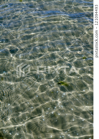 A clear water surface on the sea coast with the seashell and wavy water on a sunny day. The top view of transparent sea water at the seashore with the reflection of scattered sunlight under the water 121094493