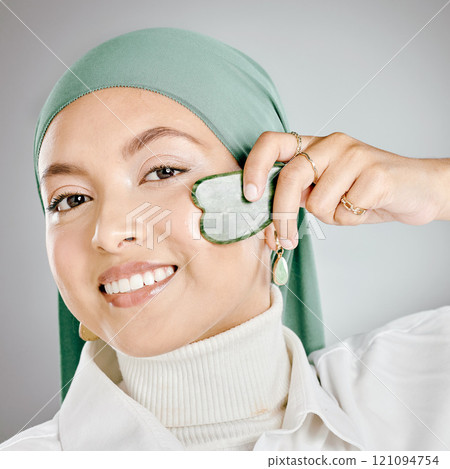 Portrait of a happy beautiful muslim woman using a gua sha on her face against a grey studio background. Young woman wearing a hijab and doing her skincare routine. Taking care of healthy skin 121094754