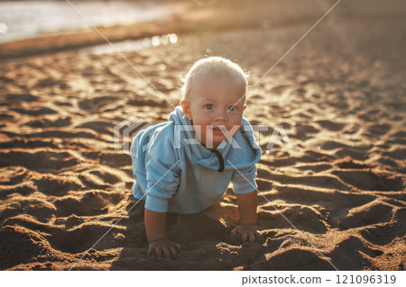Happy toddler in blue hoodie crawls along beach on sunny day 121096319