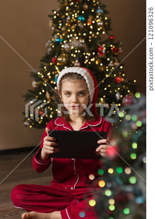 Happy girl of 7 years old in red pajamas and Santa Claus hat sitting on living room floor next to Christmas tree and communicating on tablet Happy girl of 7 years old in red pajamas and Santa Claus hat sitting on living room floor next to Christmas tree and communicating on tablet 121096326