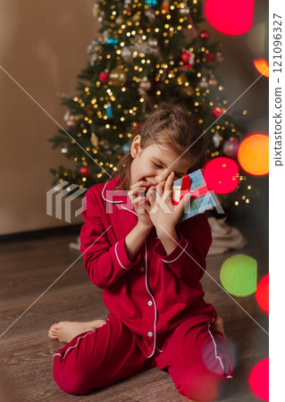 Happy girl of 7 years old in red pajamas sits on living room floor with gifts box next to Christmas tree Happy girl of 7 years old in red pajamas sits on living room floor with gifts box next to Christmas tree 121096327