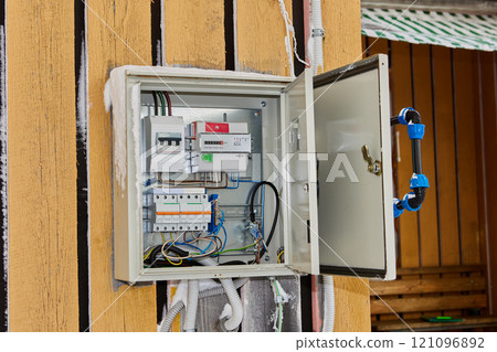 External steel electric panel with installed automatic fuses and an electrical meter is wall mounted on rural house facade. 121096892