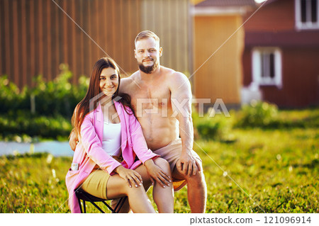 White shirtless man and woman in shorts pose for photographer on lawn near house, seated portrait of married couple in their 20s. 121096914