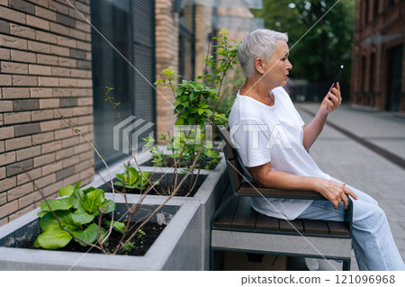 Side view of pretty mature aged female talk to friend relative child by video call outdoors. Relaxed senior woman with gray hair sitting on bench in city street and communicating using smartphone. 121096968